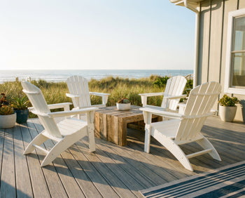 A bright, sunny coastal patio featuring a set of HDPE Adirondack chairs in crisp white arranged around a low coffee table, with beach grass and ocean visible in the background and soft morning light casting gentle shadows across a weathered composite deck