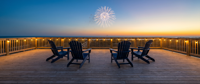 Four dark navy Adirondack chairs on a wide beachfront deck watching a fireworks burst over the ocean at sunset with warm string lights along the railing