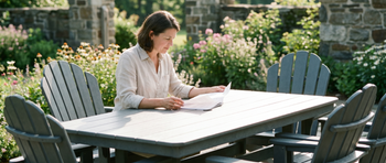 Woman reading papers at a gray outdoor dining table surrounded by Adirondack chairs in a garden patio with blooming flowers and a stone wall in the background