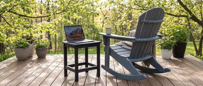 Gray Adirondack rocking chair with a draped throw blanket beside a black side table holding an open laptop and a coffee mug on a wooden deck surrounded by potted plants and budding spring trees