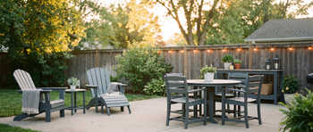 Backyard patio setup with gray Adirondack chairs, a round outdoor dining table with matching chairs, and a wooden console table strung with string lights, surrounded by a wood privacy fence and greenery at golden hour.